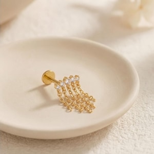 Può includere: Un orecchino a lobo dorato con retro piatto, caratterizzato da una barra orizzontale con cinque piccole pietre trasparenti a taglio rotondo. Dalla barra pendono cinque delicate catenelle dorate di varie lunghezze, ognuna con una pietra.