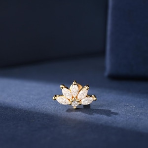 以下が含まれることがあります： 金色のスタッドピアス、花のモチーフ。 ピアスは、中央の丸いクリアな宝石の周りに配置された、6つのマーキス型のクリアな宝石が特徴です。 ピアスは濃い青色の背景に配置されています。