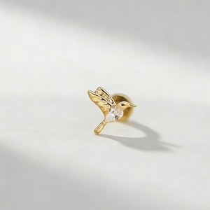 May include: A gold-colored hummingbird stud earring with a clear, sparkling gemstone on its chest. The bird is depicted in flight, with detailed wings and tail feathers. The earring is set against a soft, neutral background.