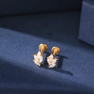 May include: A pair of gold-colored stud earrings. Each earring features a leaf design with multiple clear, sparkling stones. The earrings are set against a dark blue background, creating a contrast that highlights their details.