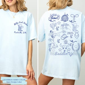 May include: Pale blue t-shirt with navy blue illustrations. Front displays "Betty Last Rodeo Nashville, TN" and a boot graphic. The back features "Yee Haw" text, a disco ball, cowboy-themed drawings, and "2025" and "Saddlin' Up" text.