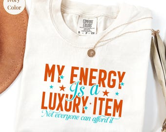 May include: Ivory-colored t-shirt with the text "MY ENERGY Is a LUXURY ITEM" in orange and teal. The shirt has a crew neck and short sleeves. A gold necklace with a pendant rests on the shirt.
