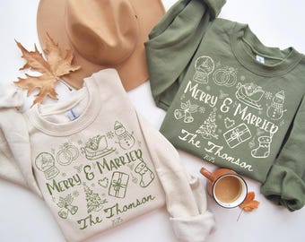 Può includere: Due felpe, una color crema e una verde oliva, con la scritta "Merry & Married" e illustrazioni a tema matrimonio. Sono visibili anche un cappello marrone e una foglia autunnale. Le felpe sono un design accogliente e festoso.