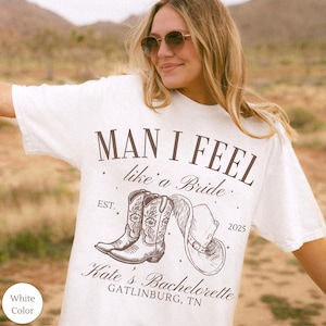 May include: White t-shirt with the text "MAN I FEEL like a Bride" and a graphic of cowboy boots and a hat. The shirt also includes "Kate's Bachelorette GATLINBURG, TN" and the year 2025.