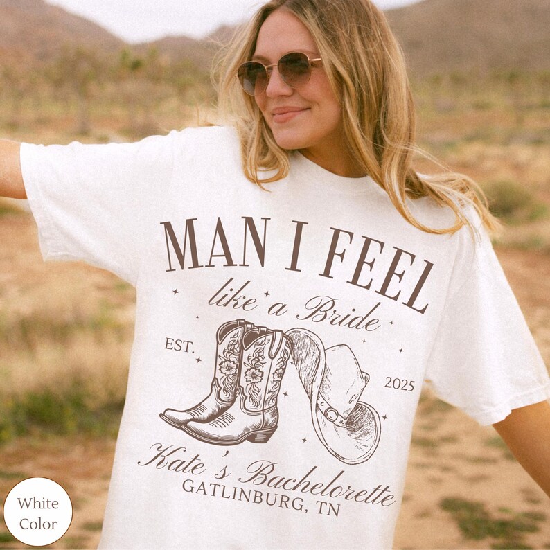 May include: White t-shirt with the text "MAN I FEEL like a Bride" and a graphic of cowboy boots and a hat. The shirt also includes "Kate's Bachelorette GATLINBURG, TN" and the year 2025.