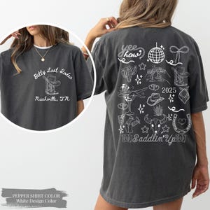 May include: Charcoal grey t-shirt with a white graphic. The front displays "Betty Last Rodeo Nashville, TN". The back has a "Yee Haw" design with cowboy-themed illustrations, the text "2025" and "Saddlin' Up".