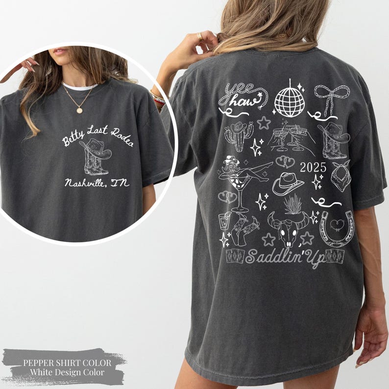May include: Charcoal grey t-shirt with a white graphic. The front displays "Betty Last Rodeo Nashville, TN". The back has a "Yee Haw" design with cowboy-themed illustrations, the text "2025" and "Saddlin' Up".