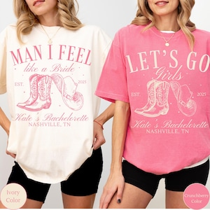May include: Two t-shirts, one ivory and one pink, with a country theme. The ivory shirt reads "Man I Feel like a Bride" and the pink shirt says "Let's Go Girls." Both shirts feature a cowboy boot and hat graphic, with "Kate's Bachelorette Nashville, TN" printed below.