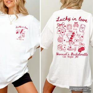 Può includere: T-shirt bianco con un design grafico rosso. La parte anteriore presenta un bicchiere da martini e il testo "Lucky in love" e "Las Vegas, NV". La parte posteriore presenta un design più grande con varie illustrazioni a tema casinò e il testo "Hannah's Bachelorette".