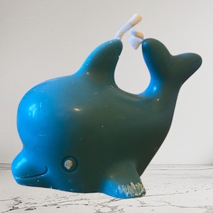 Peut inclure: Une bougie en forme de dauphin bleu et vert d'eau avec une mèche blanche. La bougie a une finition lisse et brillante et un petit détail d'œil rond. Le dauphin est posé dans une position ludique et verticale.