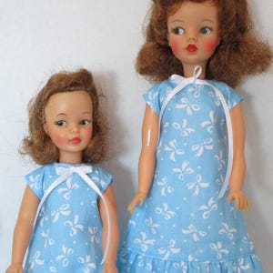 Könnte beinhalten: Zwei Vintage-Puppen, eine große und eine kleine, tragen passende hellblaue Kleider mit weißen Schleifenmustern. Die Kleider haben Rüschensaum und weiße Schleifen am Hals. Die Puppen haben rotes Haar und bemalte Gesichtszüge.