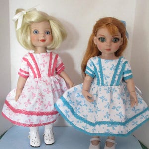 May include: Two dolls wearing white dresses with pink and blue bows. The doll on the left has blonde hair and is wearing a pink dress with white lace trim. The doll on the right has red hair and is wearing a blue dress with white lace trim.