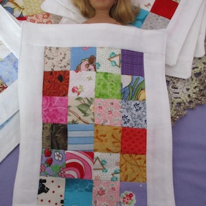 Puede incluir: Una cama de muñeca blanca con una colcha de retazos hecha de cuadrados de tela de colores. La colcha tiene un borde blanco y está llena de varios patrones y colores, incluyendo rojo, azul, verde y amarillo.