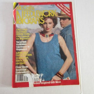 Può includere: Una copertina di rivista per McCall's Needlework & Crafts con una donna che indossa una canotta blu e un uomo che indossa un cappello da cowboy. Il testo della copertina recita "Oltre 75 fantastici progetti estivi!" e "Idee regalo ispirate al giardino".