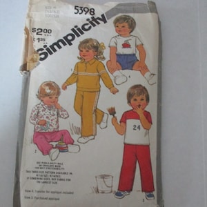 Può includere: Busta di cartamodello Simplicity vintage per abbigliamento per bambini piccoli, con illustrazioni di bambini in vari outfit. La busta è bianco sporco con il logo Simplicity e i dettagli del prodotto, compreso il prezzo.