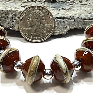 May include: A string of beads featuring glossy, dark brown, disc-shaped ceramic components with cream-colored edges, interspersed with small, reflective silver-toned spherical spacers. A United States quarter coin is placed above for scale.