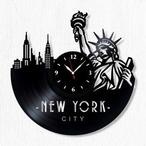 Puede incluir: Reloj de vinilo negro con una silueta del horizonte de la ciudad de Nueva York y la Estatua de la Libertad. La esfera del reloj tiene agujas negras y números blancos. El texto "NEW YORK CITY" está impreso debajo de la esfera del reloj.