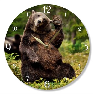 May include: A brown bear sitting in a forest setting with its paw raised in a wave. The bear is wearing a clock face with Roman numerals and gold hands. The clock face is set to 12:00.