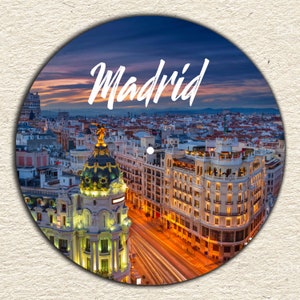 May include: An aerial view of Madrid, Spain at dusk. The city is lit up with streetlights and the sky is a vibrant blue and orange. The text "Madrid" is written in white cursive letters across the top of the image.
