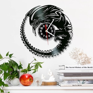 May include: A black vinyl record clock featuring a silhouette of an alien creature. The clock face is a smaller circle with black numbers and a red second hand.
