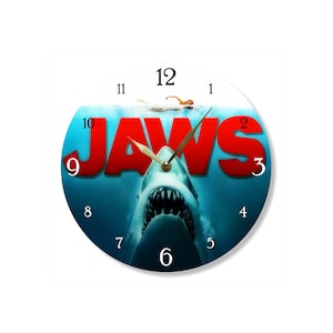 May include: A round wall clock featuring the Jaws movie poster graphic. The design displays a large shark rising from deep blue water toward a swimmer at the surface, with the red text JAWS prominently displayed. Black numbers mark the hours.