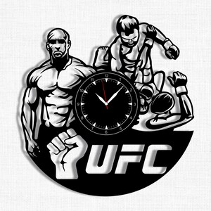 Peut inclure: Silhouette noire et blanche de deux combattants d'arts martiaux mixtes en position de combat, avec un cadran d'horloge au centre et le mot "UFC" en dessous.