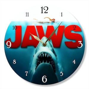 May include: A round clock with a blue and white water background. The clock face features the word "JAWS" in red letters with a shark's mouth open below. The clock hands are gold and the numbers are black.