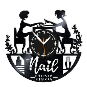 Può includere: Orologio in vinile nero a tema nail studio. L'orologio presenta sagome di due donne, una delle quali fa una manicure. Il quadrante è nero con numeri e lancette bianche. Le parole "Nail Studio" sono scritte in bianco.