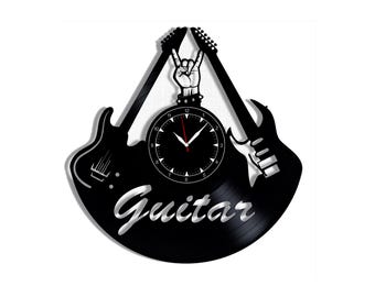 Könnte beinhalten: Eine schwarze Wanduhr aus einer Schallplatte mit zwei nach oben gerichteten E-Gitarren-Silhouetten. Eine Hand mit Rock-Geste befindet sich zwischen den Gitarrenhälsen über einem runden Zifferblatt mit weißen Zahlen. Das Wort Guitar ist in Silber gehalten.