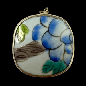 May include: A round porcelain pendant with a gold-tone metal frame. The pendant features a blue and green floral design on a white background.