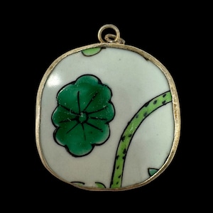 Peut inclure: Un pendentif en argent avec un charme ovale en porcelaine blanche orné d'un motif floral vert. Le charme a une fleur verte avec un contour noir et une tige verte avec des points noirs.