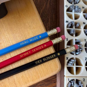May include: Three pencils with erasers and text: "HERO MODE", "SPIDEY SENSES", and "WEB SLINGER". The pencils are blue, red, and black, respectively. A wooden block and a box with metal letter stamps are in the background.