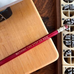 Könnte beinhalten: Ein roter Bleistift mit der Aufschrift "NOTHING CAN STOP US NOW" liegt auf einer Holzoberfläche. Daneben befinden sich ein Holzblock und eine Box mit Metallbuchstaben. Der Bleistift hat einen rosa Radiergummi.