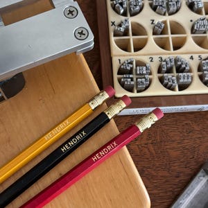May include: Three pencils in yellow, black, and red, each with "HENDRIX" printed on them. The pencils have gold-colored metal bands and pink erasers. A wooden block and a metal tool are also visible.