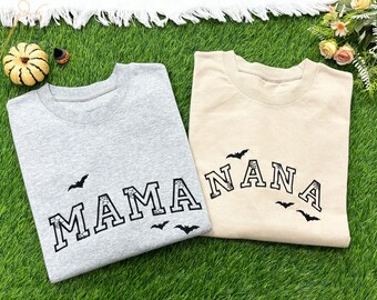 May include: Two crewneck sweatshirts, one gray and one beige, with the words "MAMA" and "NANA" in black, arched lettering. Each shirt has black bat graphics. A small pumpkin and a decorative item are visible.