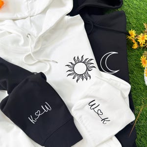 Può includere: Felpe con cappuccio bianche e nere con grafiche di sole e luna. La felpa bianca ha un design a forma di sole e la felpa nera ha una luna crescente. Le maniche presentano le iniziali con un cuore. Un set coordinato e accogliente.