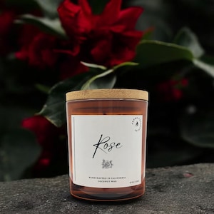 May include: A brown glass candle jar with a wooden lid. The label reads "Rose" and "Handcrafted in California Coconut Wax".