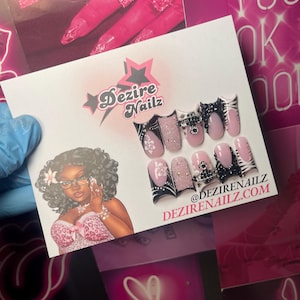 May include: A card featuring a set of artificial nails with various designs, including pink, black, and glitter accents. The card also displays the "Dezire Nailz" logo and a cartoon illustration of a woman.