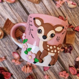Puede incluir: Una taza de cerámica rosa con un diseño de ciervo en 3D. El ciervo es marrón con manchas blancas y tiene una nariz rosa. La taza tiene un borde y un asa blancos.
