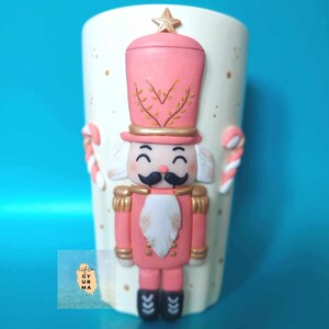 May include: A white ceramic mug with a pink and gold nutcracker design. The nutcracker is wearing a red and gold uniform and has a white beard. The mug is decorated with gold glitter and candy cane accents.