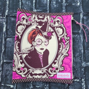 May include: A pink zippered pouch with a black and white illustration of a girl wearing a hat and glasses. The girl is holding a bird in her hand. The pouch has a pink and white floral pattern and the text "pinkbody" in white.