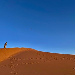 Puede incluir: Un paisaje desértico con una duna de arena bajo un cielo azul claro. Una persona está de pie en la cima de la duna, con huellas en la arena. La luna es visible.