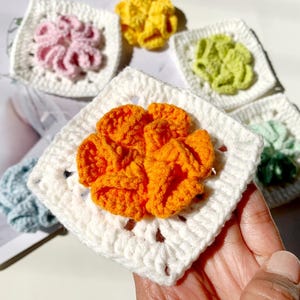 May include: Five crocheted squares with different colored flowers in the center. The flowers are orange, pink, yellow, green, and blue. The squares are white.