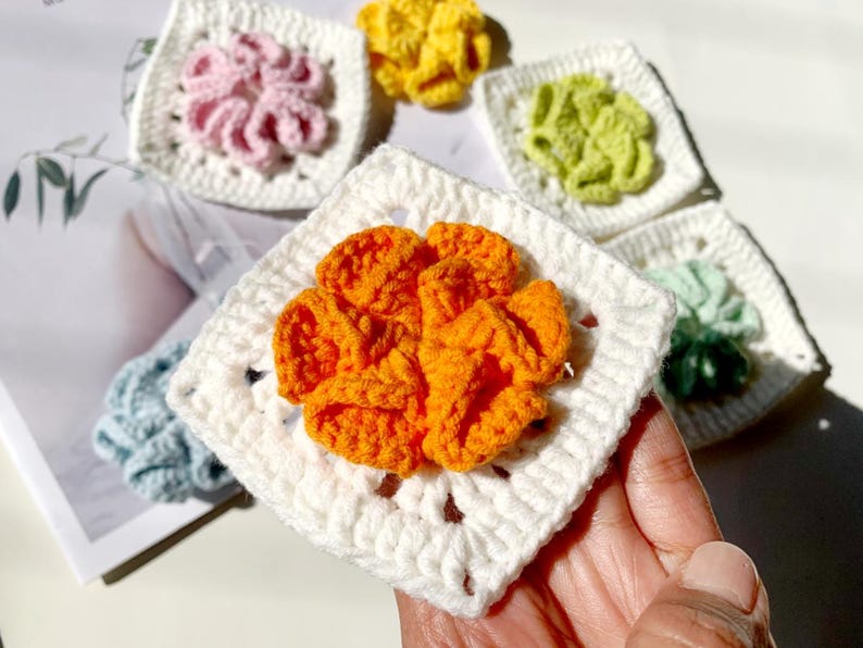 May include: Five crocheted squares with different colored flowers in the center. The flowers are orange, pink, yellow, green, and blue. The squares are white.