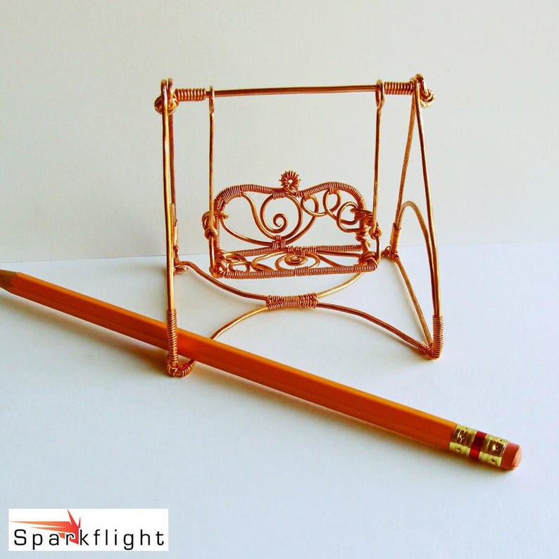 May include: A miniature copper wire swing with a decorative back, next to a standard orange pencil. The swing is made of thin copper wire, with intricate details in the seat's design. The pencil is orange with a gold band and red eraser.