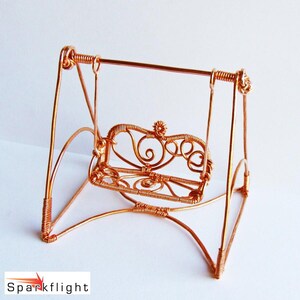 May include: A handcrafted copper wire swing with an ornate seat design. The swing features a curved frame and a horizontal bar at the top. The seat has a decorative, scrolled pattern and a small floral accent. The overall design is delicate and artistic.