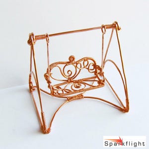 May include: A miniature copper-colored wire swing with a decorative back and seat. The swing has a simple frame and is suspended from a horizontal bar. The design includes swirls and a floral-like detail on the backrest. The base is a simple A-frame.