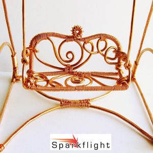 May include: A handcrafted, miniature copper wire chair with intricate details. The chair features a decorative back with swirls and a small flower-like design at the top. The legs and frame are made of copper wire. The chair is a decorative item.