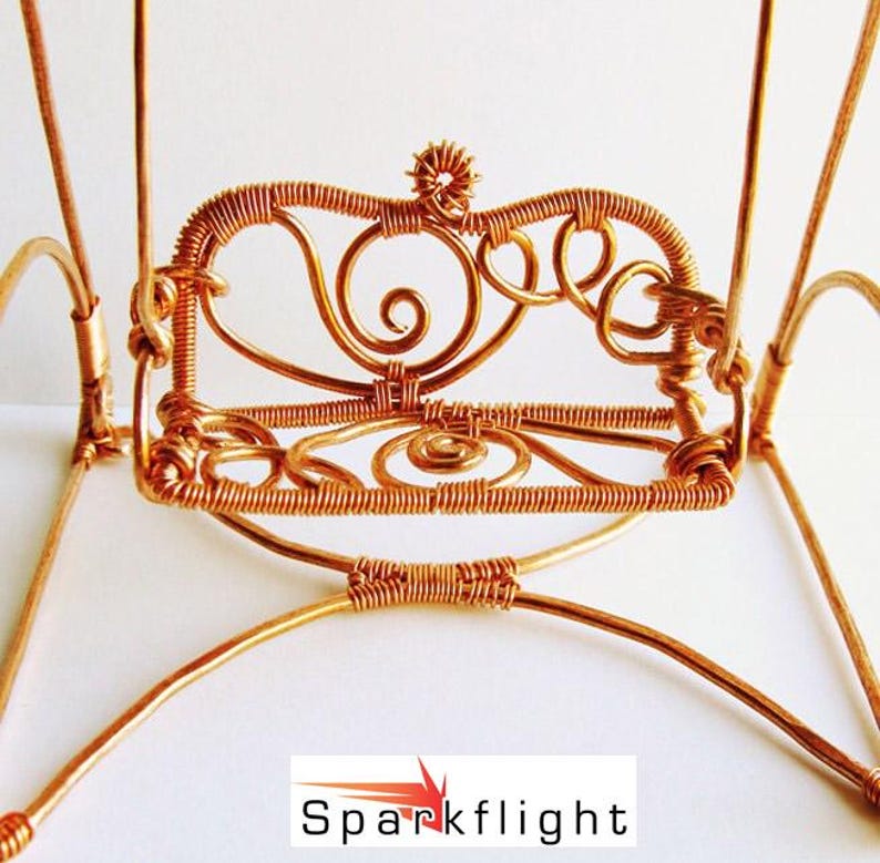 May include: A handcrafted, miniature copper wire chair with intricate details. The chair features a decorative back with swirls and a small flower-like design at the top. The legs and frame are made of copper wire. The chair is a decorative item.