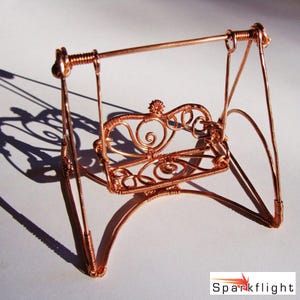 May include: A miniature copper wire swing with an ornate, heart-shaped backrest. The swing features intricate wire detailing and a decorative floral element at the top. The design includes curved supports and a horizontal bar for hanging.